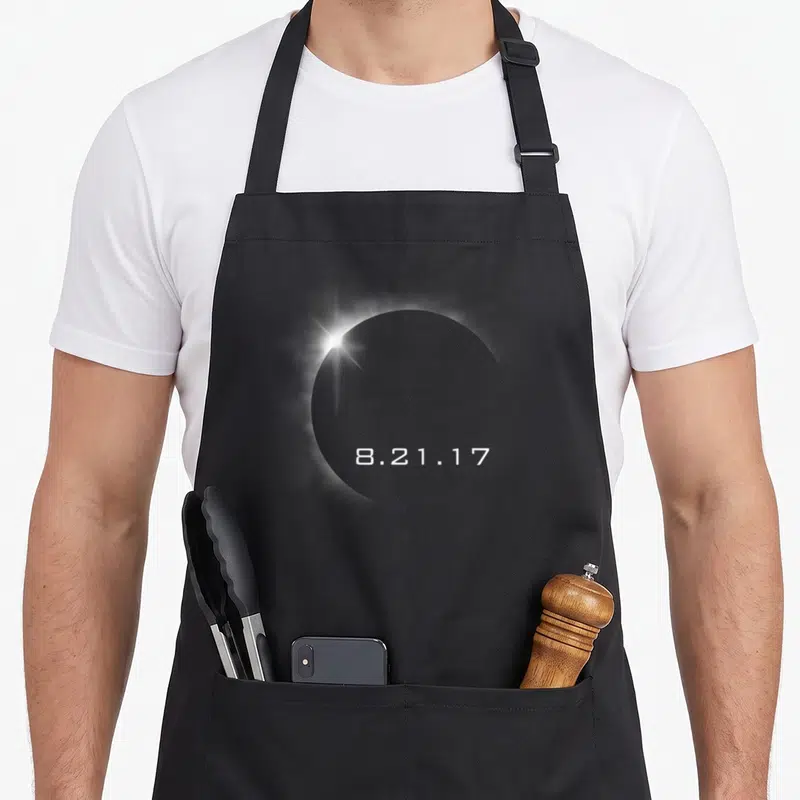 Total summer aprons - White 8.21.17 text below a bright diamond ring light on a grey circular shape — shown on a black cotton canvas apron against a white background