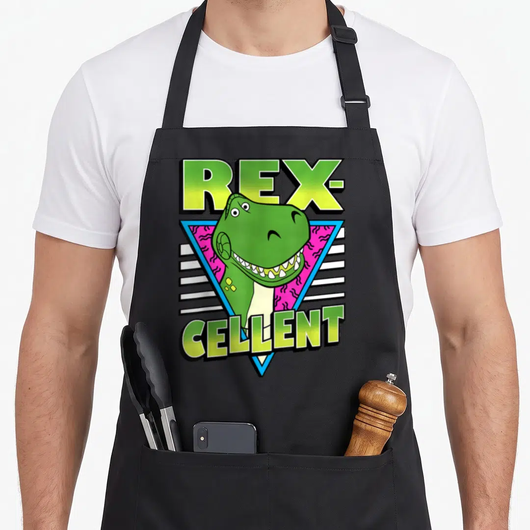 Rex dog breed aprons - REX-CELLENT pun with green T-Rex head in blue triangle on pink zigzag background — shown on a black cotton canvas apron against a white background