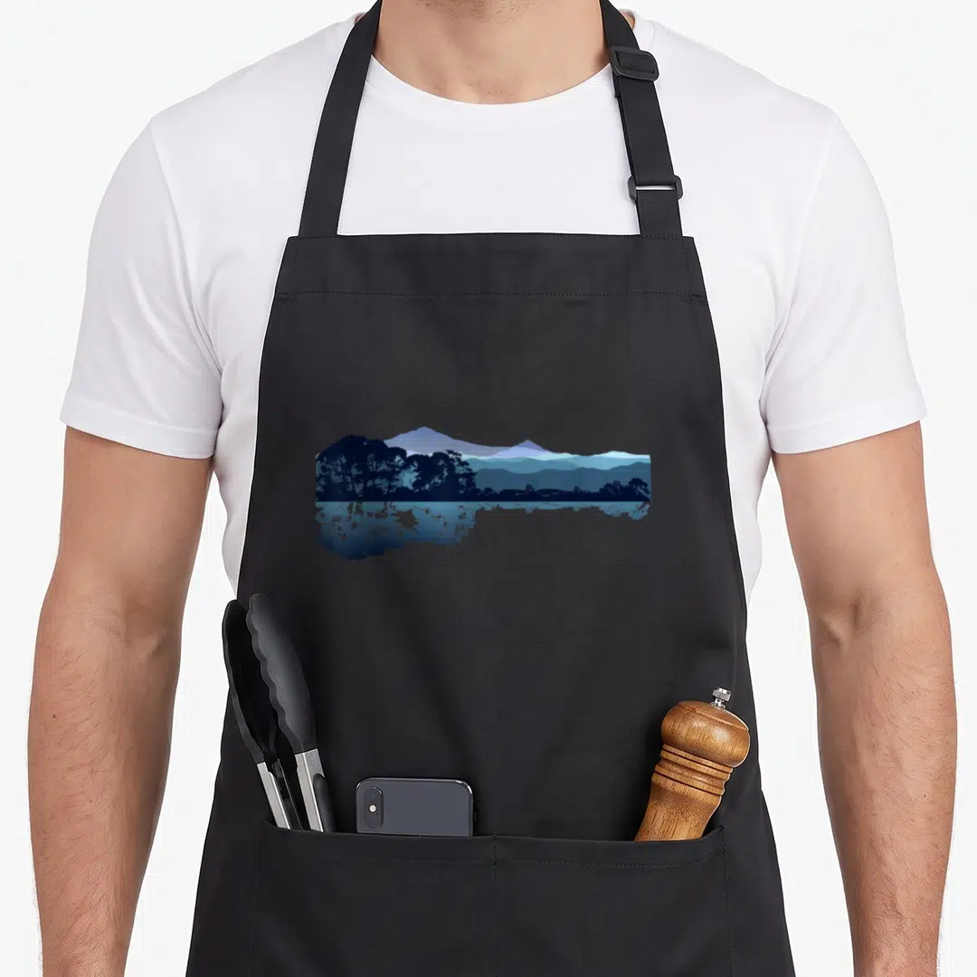 Nature decorating apron - Minimalist blue landscape with mountains and trees reflected in water — shown on a black cotton canvas apron against a white background
