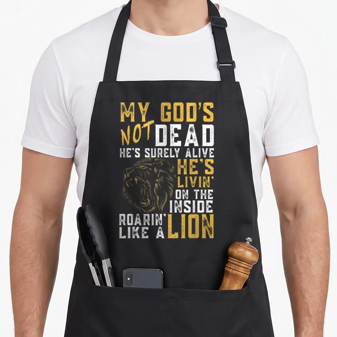 God cross apron - Yellow lion head illustration with stacked white and gold text reading MY GOD'S NOT DEAD HE'S SURELY — shown on a black cotton canvas apron against a white background