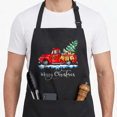 Truck old fashioned aprons - Red vintage truck carrying Christmas tree and gifts with JOY banner and Merry Christmas — shown on a black cotton canvas apron against a white background