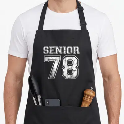 Reunion senior apron - White distressed SENIOR 78 text stacked on grey background — shown on a black cotton canvas apron against a white background
