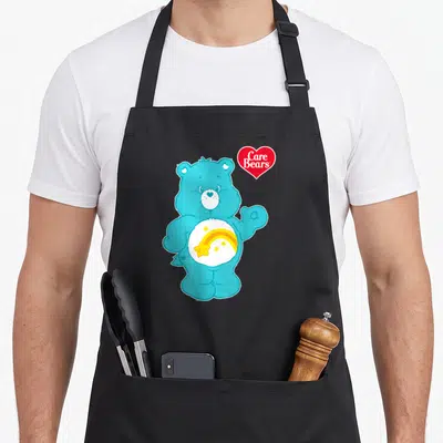 Wish care apron - Blue cartoon bear with rainbow and stars on belly next to red heart logo — shown on a black cotton canvas apron against a white background