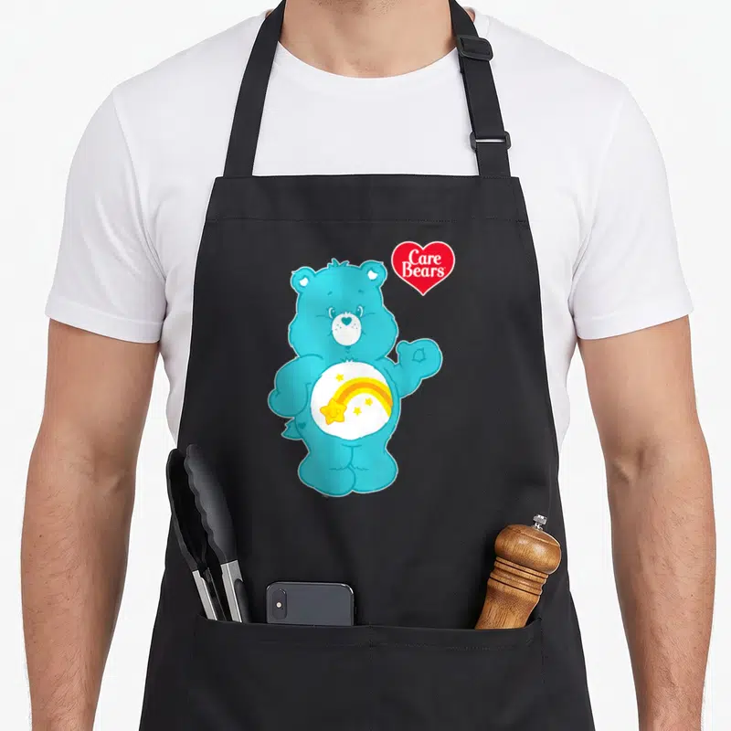 Wish care apron - Blue cartoon bear with rainbow and stars on belly next to red heart logo — shown on a black cotton canvas apron against a white background