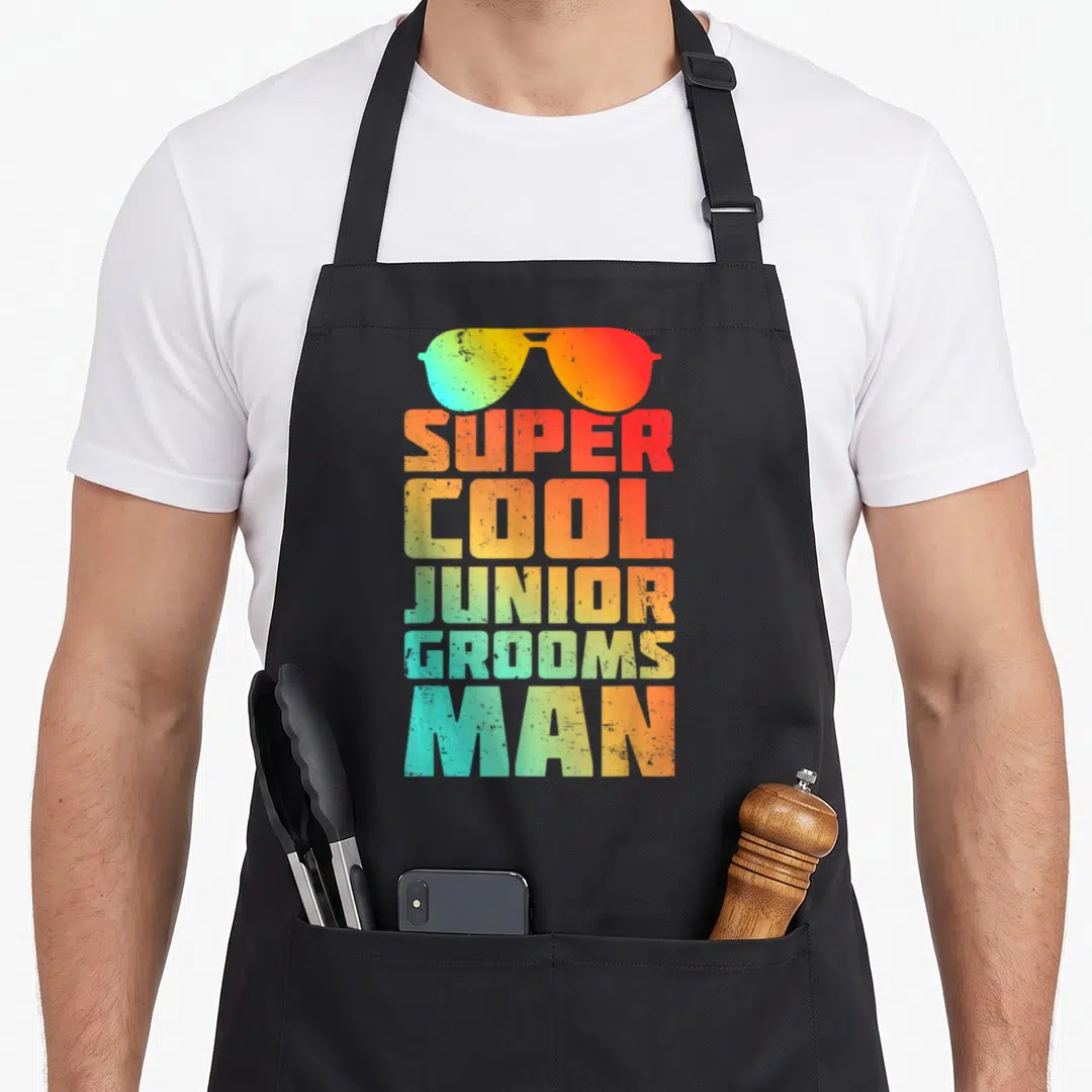 Wedding junior apron - Retro sunglasses above stacked SUPER COOL JUNIOR GROOMSMAN text in gradient colors — shown on a black cotton canvas apron against a white background