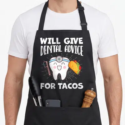 Advice dentist apron - Cartoon tooth holding toothbrush and taco with text WILL GIVE DENTAL ADVICE FOR TACOS — shown on a black cotton canvas apron against a white background