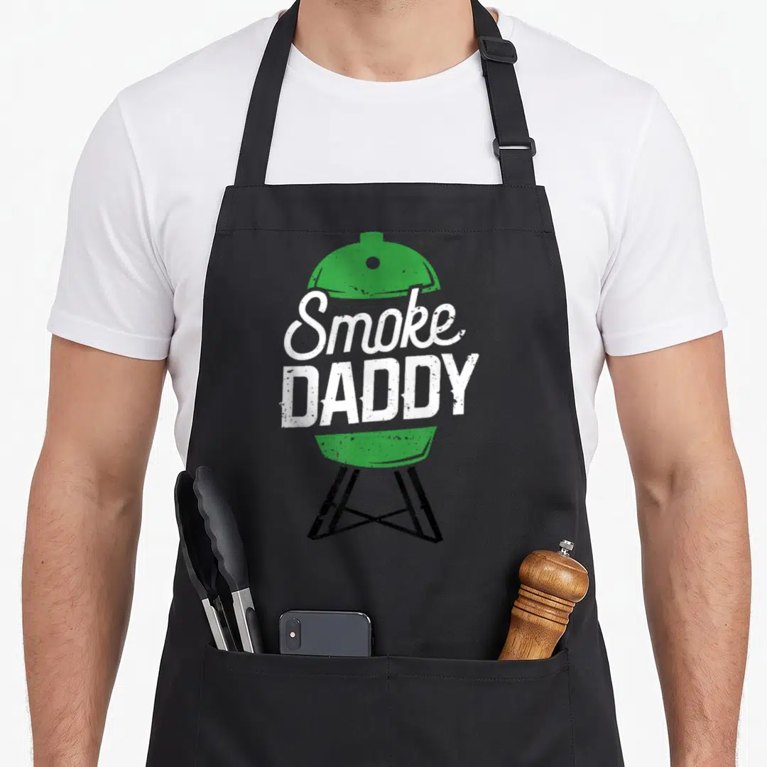 Smoke egg apron - Green and black cartoon smoker grill with white Smoke DADDY text overlay — shown on a black cotton canvas apron against a white background