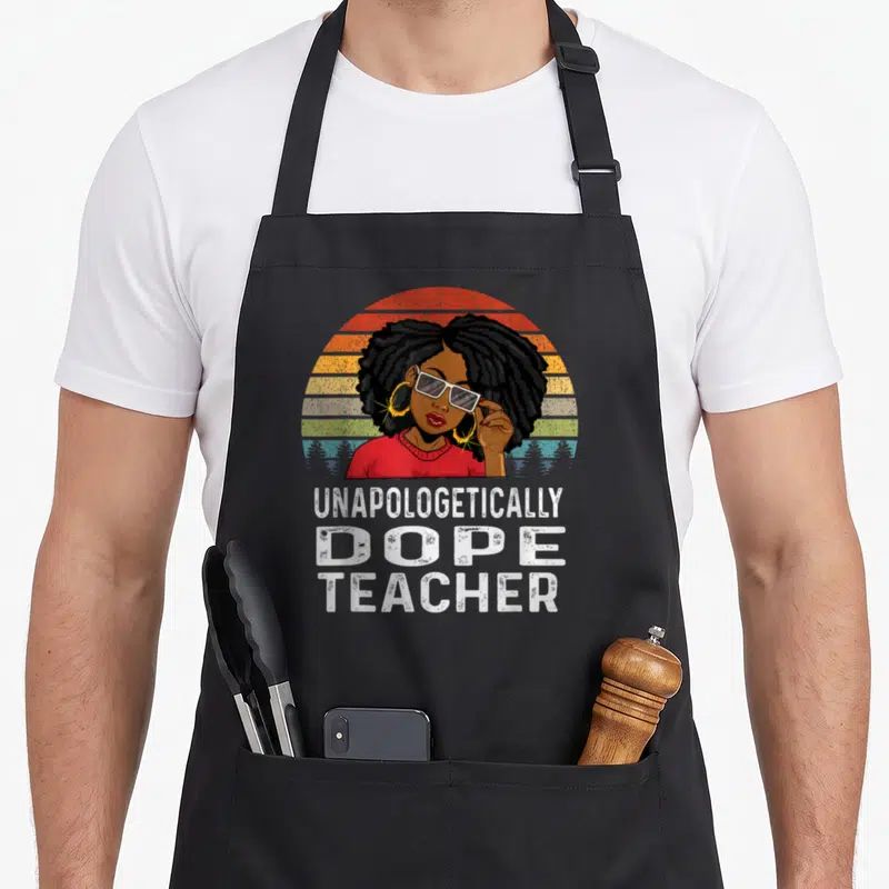 Unapologetically teacher apron - Woman with afro and glasses in front of red orange yellow striped sun with stacked — shown on a black cotton canvas apron against a white background