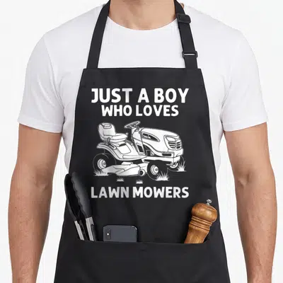Lawn farm apron - White line art riding mower with text JUST A BOY WHO LOVES LAWN MOWERS on grass patches — shown on a black cotton canvas apron against a white background