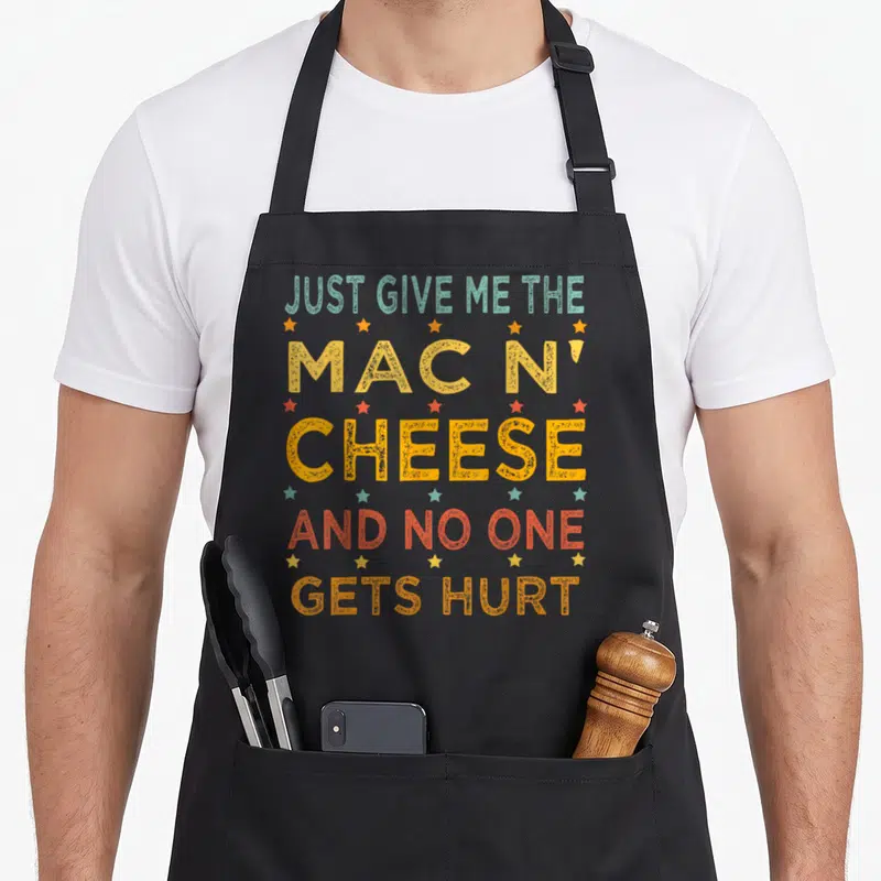 Christmas mac apron - Stacked distressed text Just Give Me The Mac N' Cheese And No One Gets Hurt with yellow red teal — shown on a black cotton canvas apron against a white background