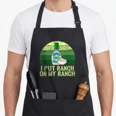Funny ranch dressing apron - Ranch dressing bottle and bowl on green striped background with pun text — shown on a black cotton canvas apron against a white background