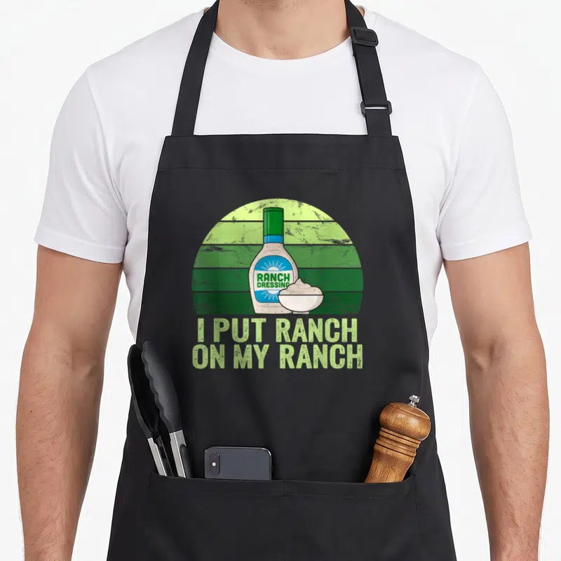 Funny ranch dressing apron - Ranch dressing bottle and bowl on green striped background with pun text — shown on a black cotton canvas apron against a white background