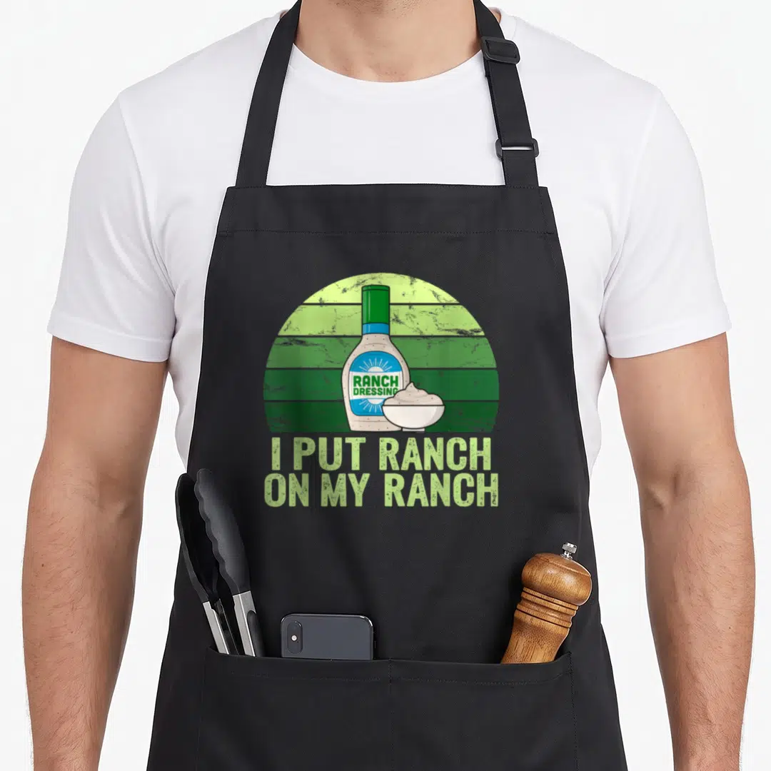 Funny ranch dressing apron - Ranch dressing bottle and bowl on green striped background with pun text — shown on a black cotton canvas apron against a white background
