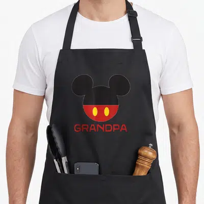 Icon father day apron - Stylized Mickey Mouse head in black red and yellow above red GRANDPA text — shown on a black cotton canvas apron against a white background
