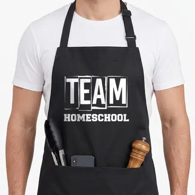 Homeschooler cute teacher apron - White text reading TEAM inside four blocks above HOMESCHOOL on gray background — shown on a black cotton canvas apron against a white background