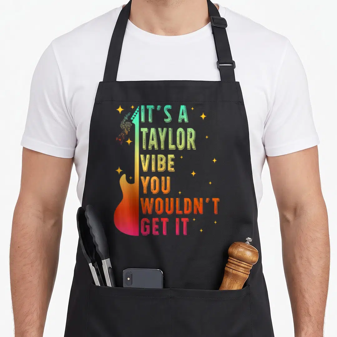 Taylor novelty apron - Vertical guitar with rainbow gradient and stacked text reading IT'S A TAYLOR VIBE YOU WOULDN'T — shown on a black cotton canvas apron against a white background