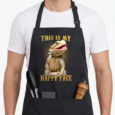 Bearded face apron - Photo of bearded dragon with THIS IS MY HAPPY FACE text and orange halftone dots — shown on a black cotton canvas apron against a white background