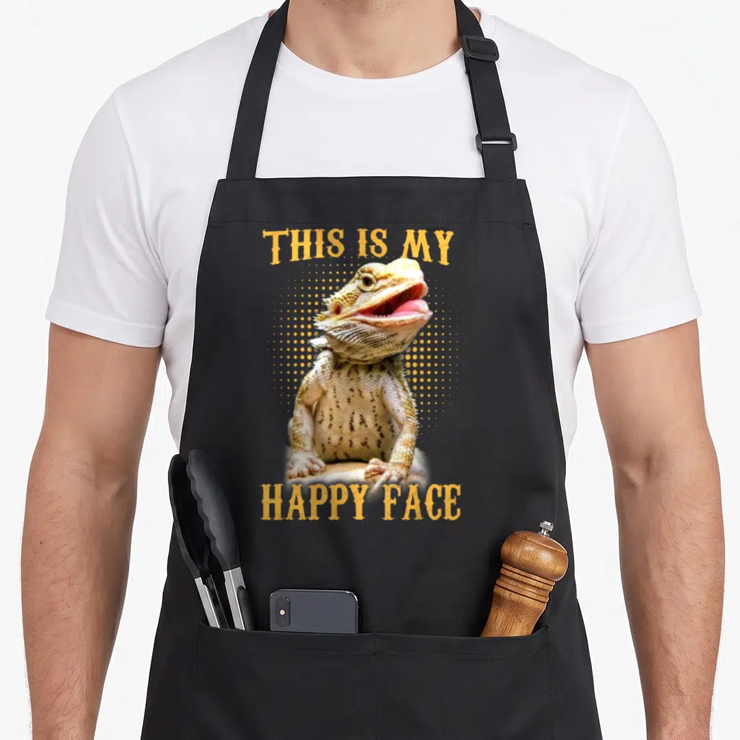 Bearded face apron - Photo of bearded dragon with THIS IS MY HAPPY FACE text and orange halftone dots — shown on a black cotton canvas apron against a white background
