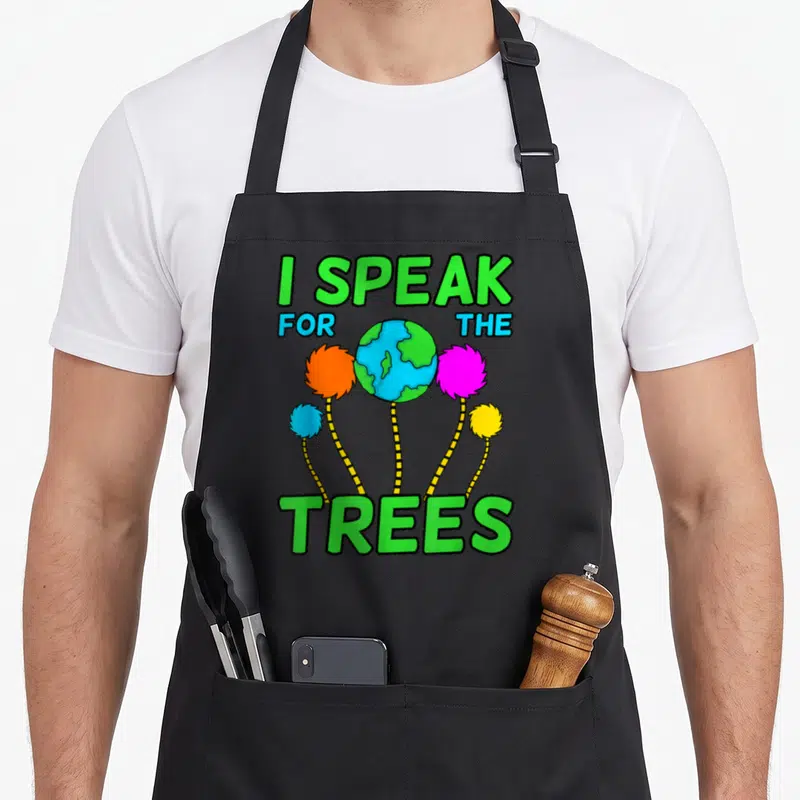 Planet tree apron - I SPEAK FOR THE TREES text around cartoon earth and four colorful fluffy trees — shown on a black cotton canvas apron against a white background