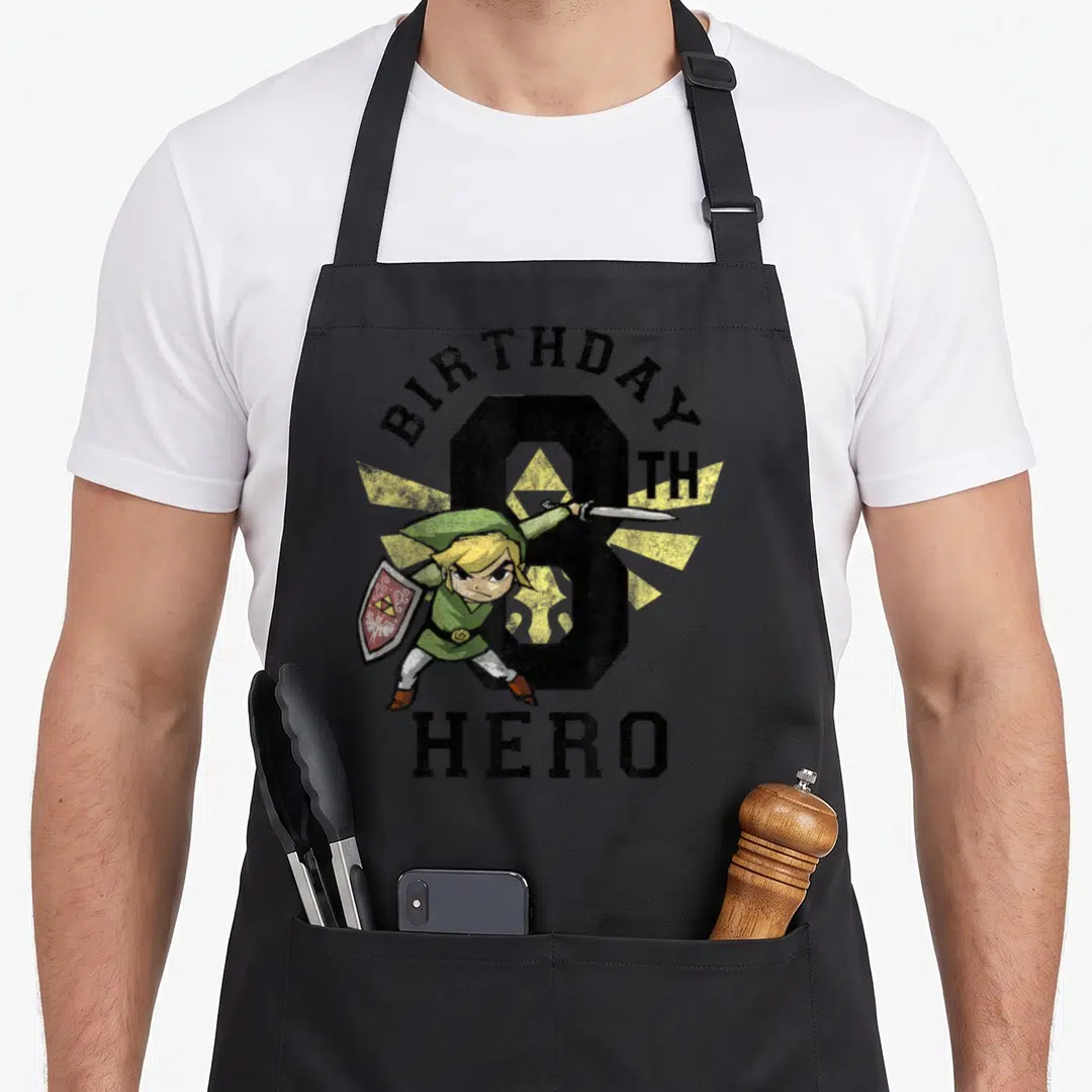 Hero birthday apron - Cartoon Link with sword and shield in front of distressed number 8 and yellow sunburst — shown on a black cotton canvas apron against a white background