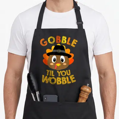 Gobble thanksgiving apron - Cartoon turkey head in black hat with GOBBLE TIL YOU WOBBLE text in orange and yellow — shown on a black cotton canvas apron against a white background