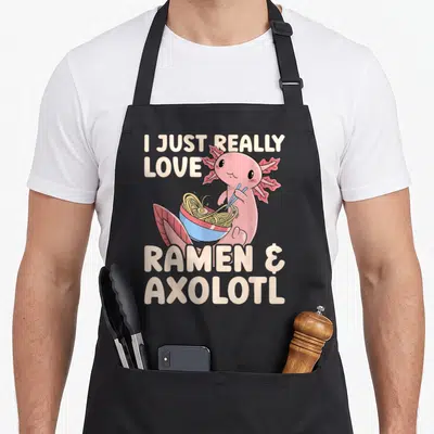 Japanese i love ramen apron - Pink axolotl holding blue ramen bowl with yellow noodles and split text — shown on a black cotton canvas apron against a white background