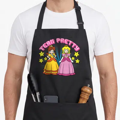 Peach daisy apron - TEAM PRETTY text over two princesses standing side-by-side with yellow stars and blue rays — shown on a black cotton canvas apron against a white background