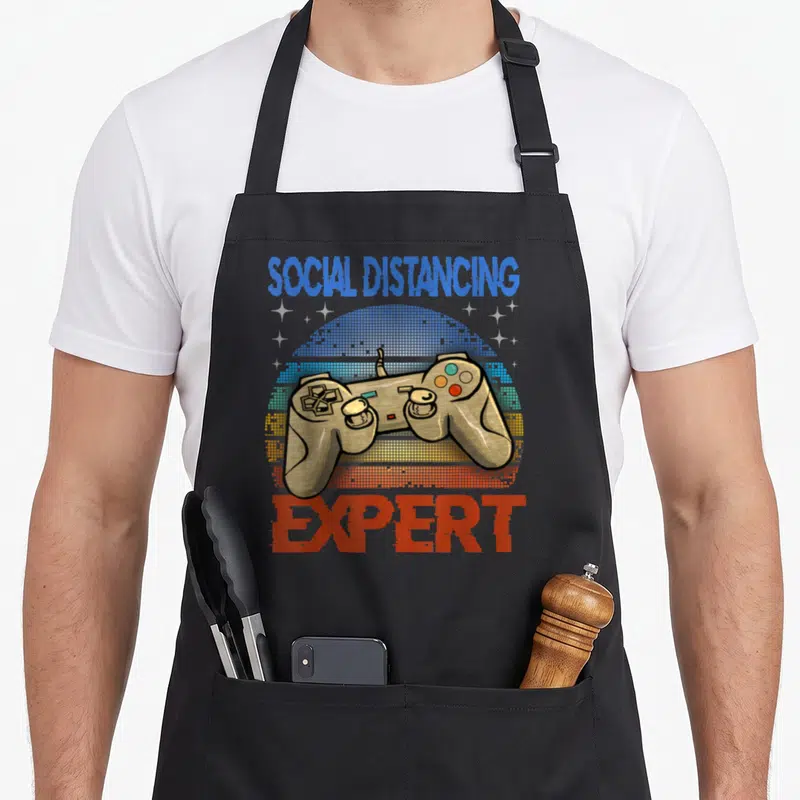 Distancing gaming apron - Retro game controller on striped sun with Social Distancing Expert text — shown on a black cotton canvas apron against a white background