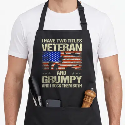 Grumpy grandpa apron - Distressed USA flag with soldier, helicopter, tank silhouettes and stacked text about veteran — shown on a black cotton canvas apron against a white background