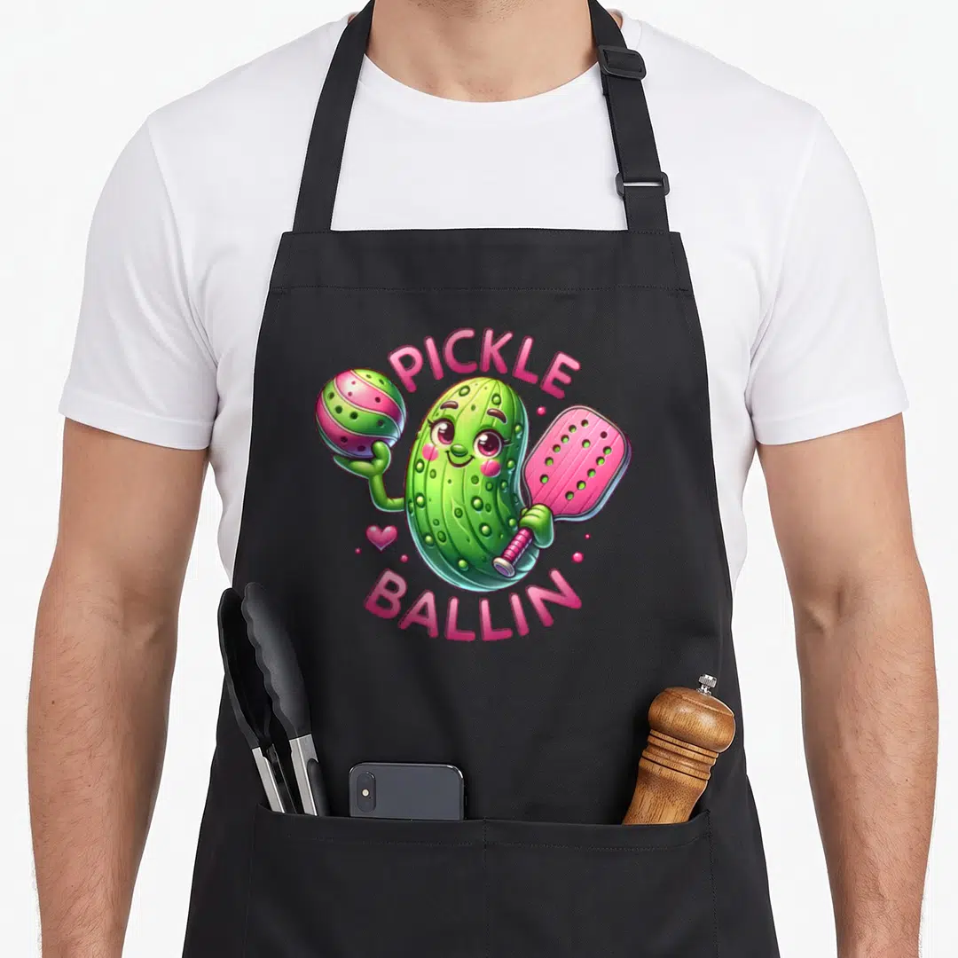 Ballin pickle apron - Cartoon green pickle holding pink paddle and striped ball with arched pink text — shown on a black cotton canvas apron against a white background