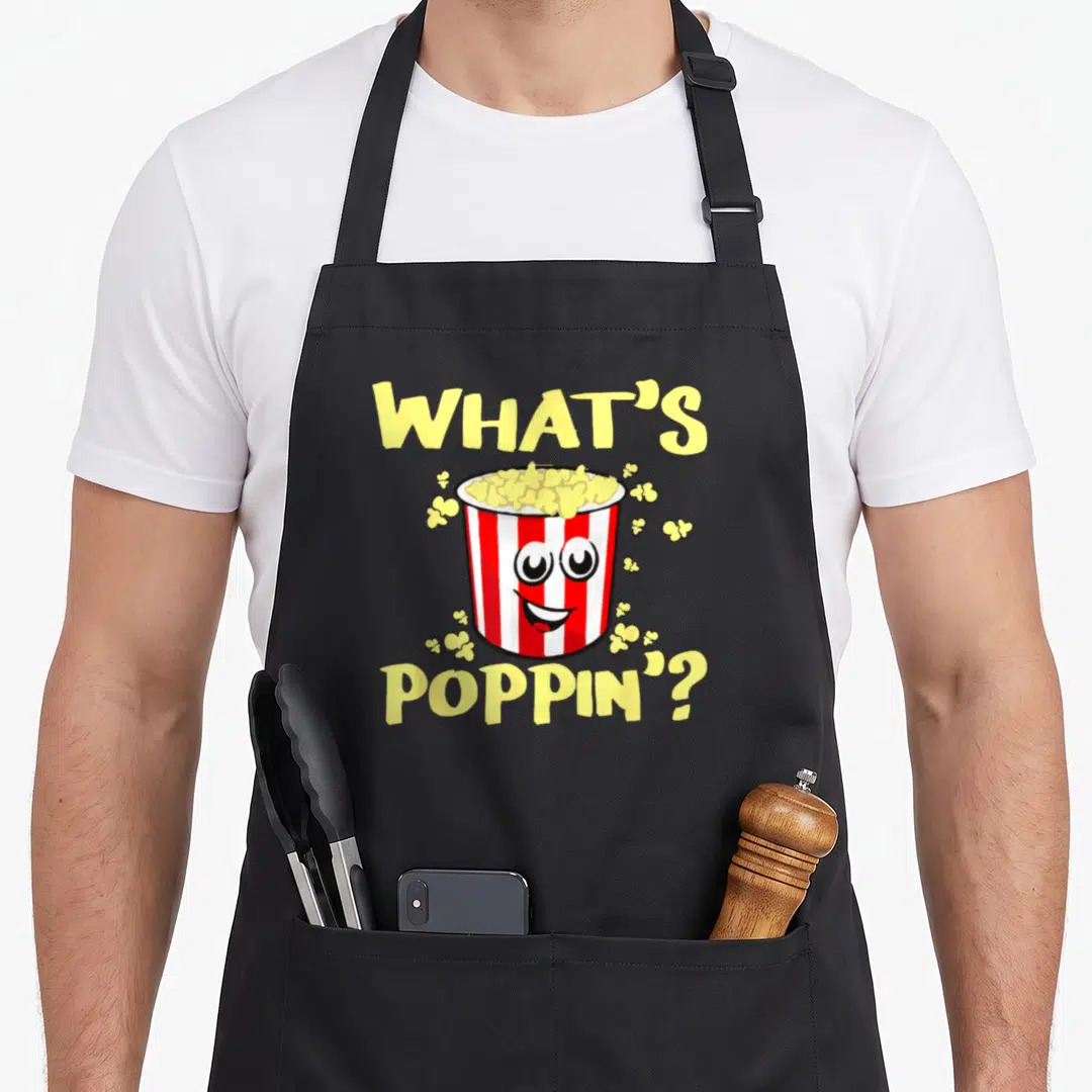 Poppin funny popcorn apron - Cartoon popcorn bucket with face and yellow text what's poppin — shown on a black cotton canvas apron against a white background