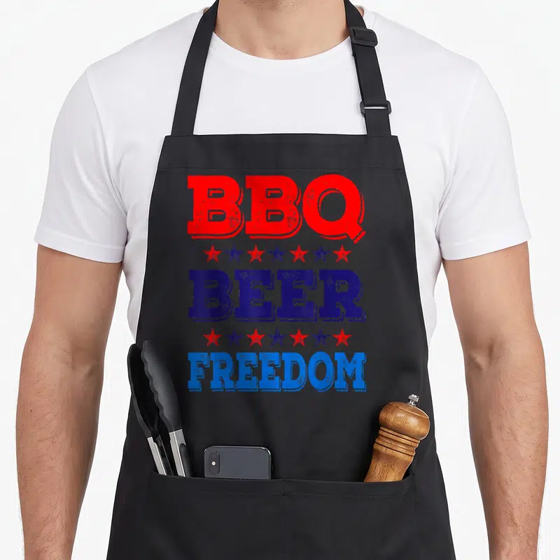 Summer 4th of july apron - Stacked red and blue text reading BBQ BEER FREEDOM separated by alternating star rows — shown on a black cotton canvas apron against a white background