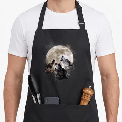 Three wolves apron - Three wolf heads howling at a full moon with stars in grey, white, and brown — shown on a black cotton canvas apron against a white background