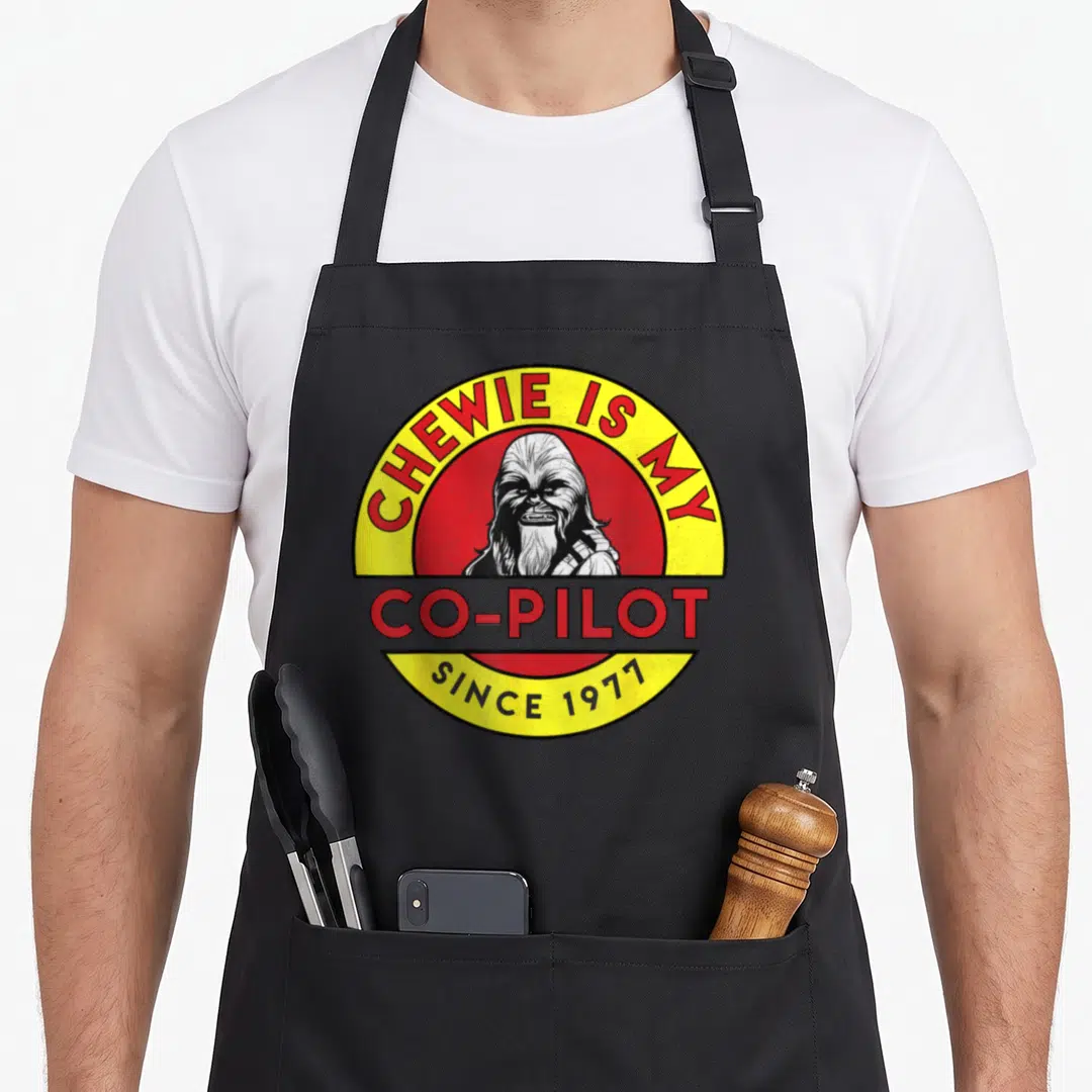 Chewie chef and co aprons - Retro red and yellow circle with Chewbacca portrait and co-pilot text — shown on a black cotton canvas apron against a white background