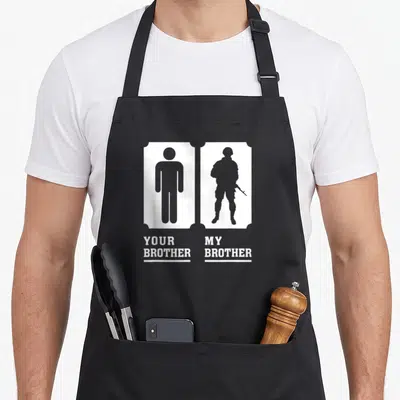 Brother military apron - YOUR BROTHER MY BROTHER text below grey silhouettes of a civilian and a soldier in white — shown on a black cotton canvas apron against a white background