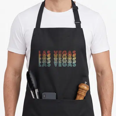 Las 70s apron - Stacked Las Vegas text in red yellow beige and blue dotted marquee font — shown on a black cotton canvas apron against a white background