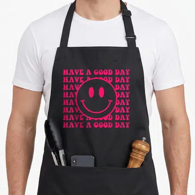 Trendy face on apron - Pink smiley face centered over eight stacked lines of text reading HAVE A GOOD DAY — shown on a black cotton canvas apron against a white background