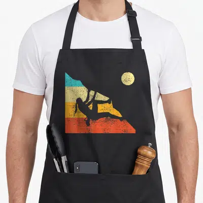 Free bouldering apron - Retro white climber silhouette against blue yellow orange and red mountain bands with beige sun — shown on a black cotton canvas apron against a white background