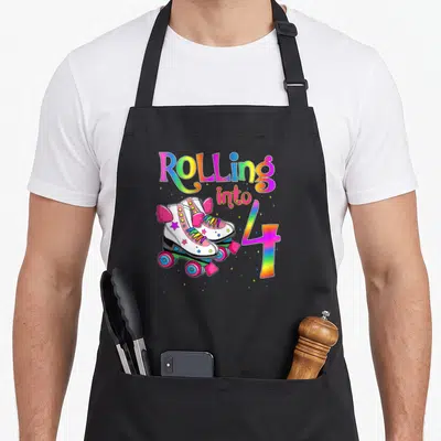 Funny rolling turning apron - Rainbow Rolling into 4 text with pink winged roller skates and scattered stars in cartoon — shown on a black cotton canvas apron against a white background