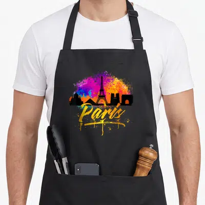 Skyline paris apron - Black Paris skyline silhouette over pink, blue, and orange paint splatter with yellow script text — shown on a black cotton canvas apron against a white background