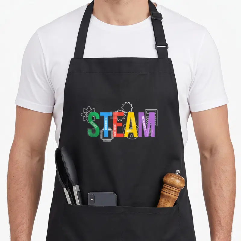 Letter teacher apron - Colorful STEAM acronym letters with vertical subtext and white atom gear chip palette calculator — shown on a black cotton canvas apron against a white background