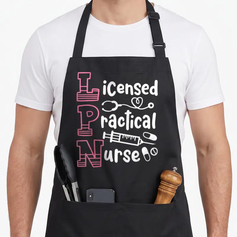 Funny lpn apron - Stacked pink LPN letters with white stethoscope syringe and pills typography design — shown on a black cotton canvas apron against a white background