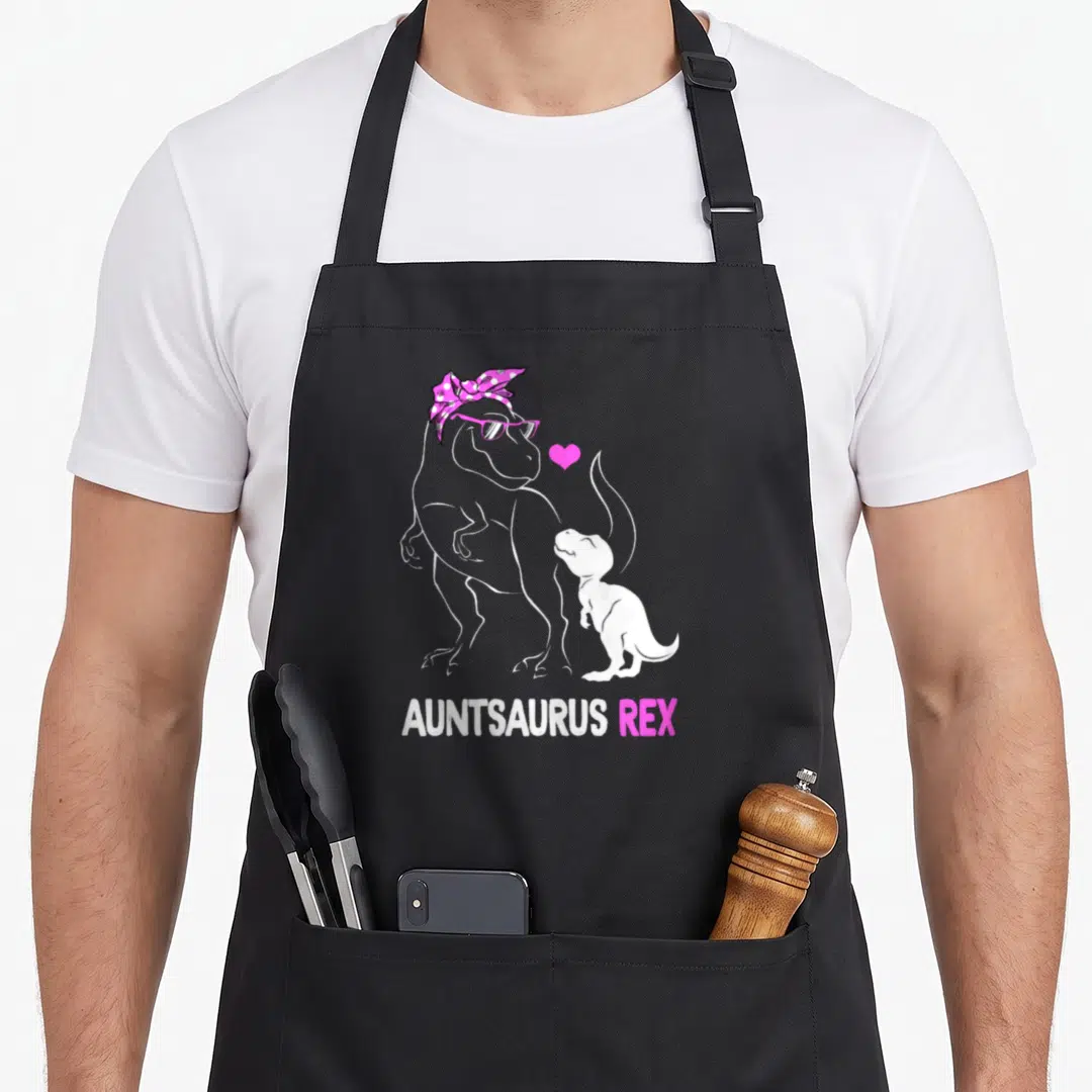 Auntie dinosaur apron - White T-Rex with pink bandana and glasses beside baby T-Rex and pink heart, Auntsaurus Rex text — shown on a black cotton canvas apron against a white background