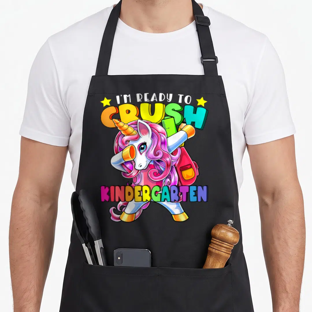 School unicorn aprons - Dabbing unicorn with pink mane, backpack, ruler, and colorful text ready to crush kindergarten — shown on a black cotton canvas apron against a white background