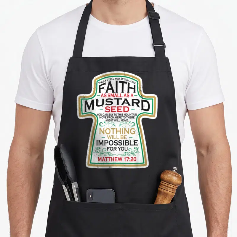 Mustard christian apron - Matthew 17:20 typography inside white cross with green border and gold trim — shown on a black cotton canvas apron against a white background
