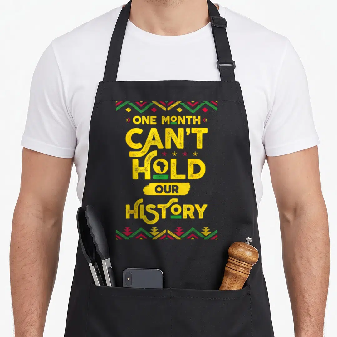Month african apron - Stacked yellow text ONE MONTH CAN'T HOLD OUR HISTORY with Africa map in O and geometric borders — shown on a black cotton canvas apron against a white background