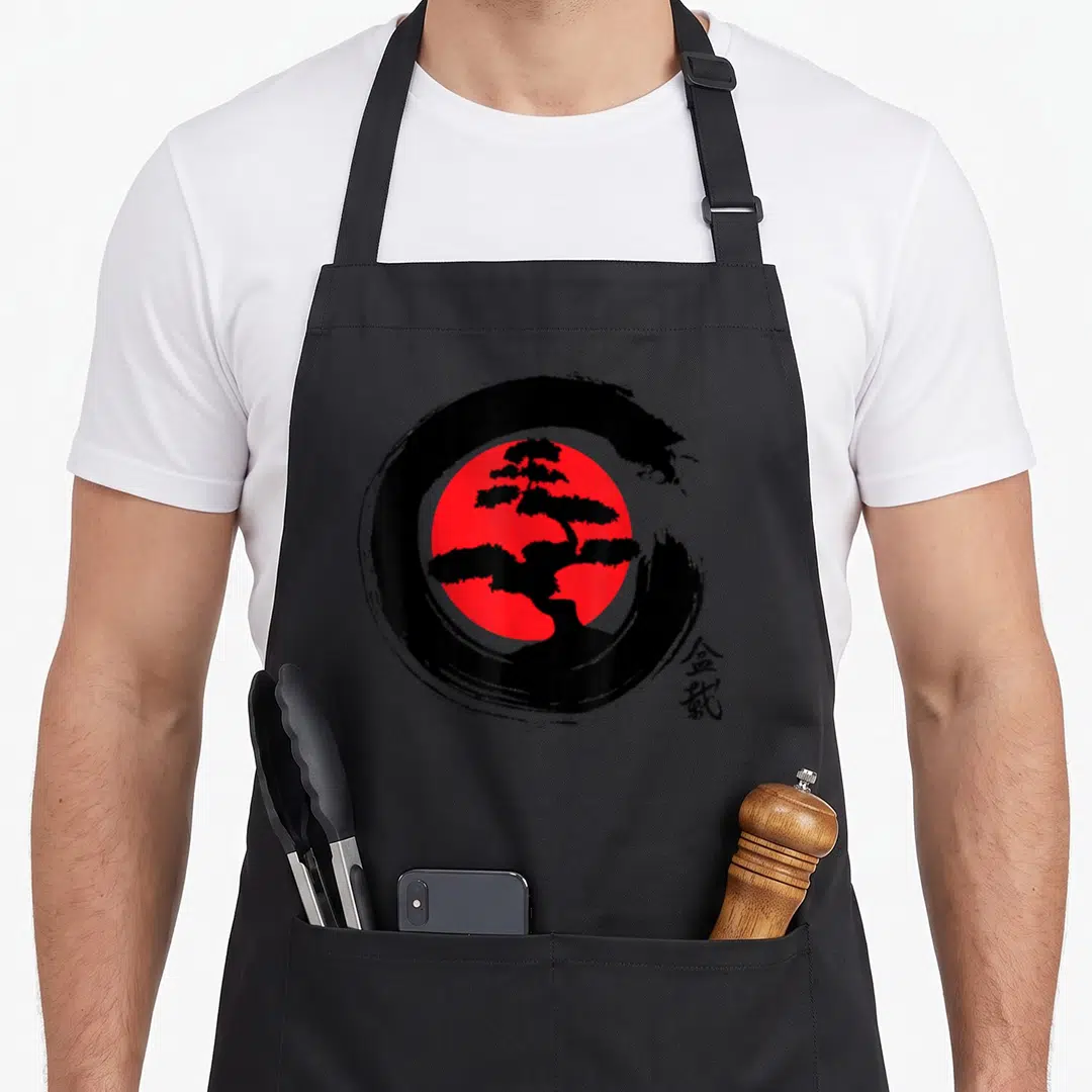 Calligraphy tree apron - Black bonsai silhouette on red sun with black brush circle and kanji characters — shown on a black cotton canvas apron against a white background