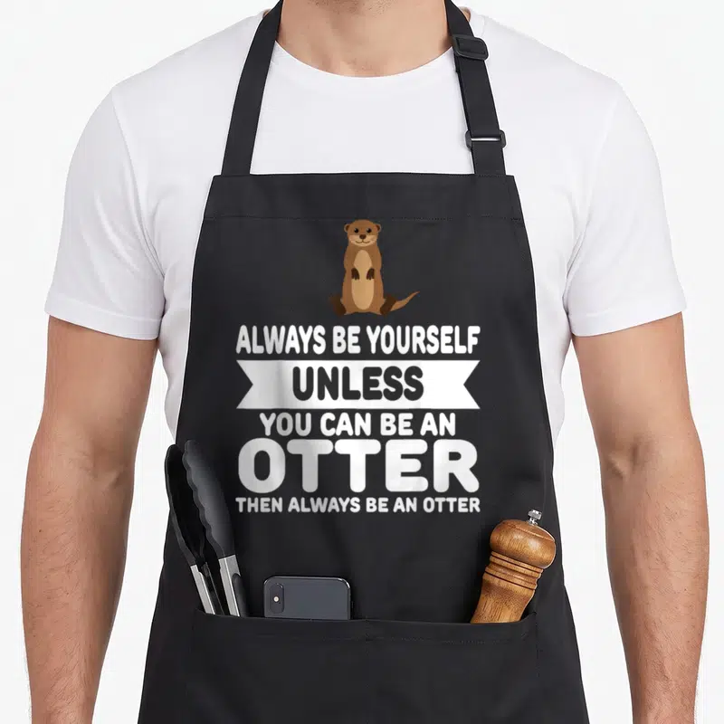 Alway unless you apron - Brown cartoon otter sitting above white text reading ALWAYS BE YOURSELF UNLESS YOU CAN BE — shown on a black cotton canvas apron against a white background