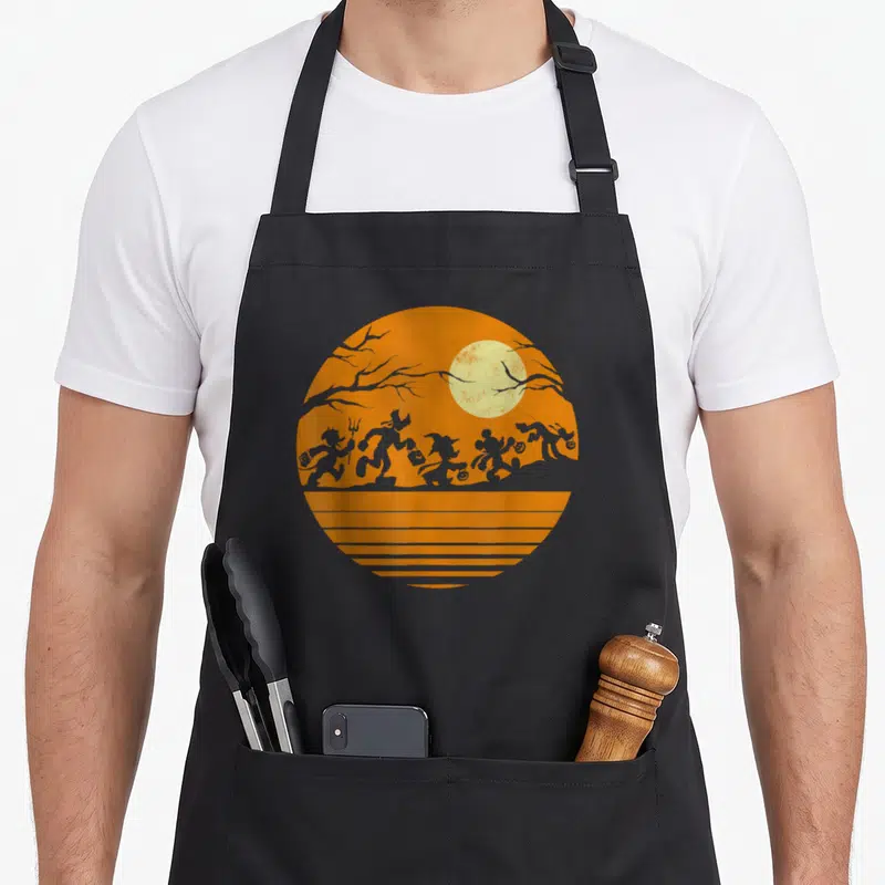 Friend halloween apron - Circular orange graphic with grey silhouettes, pale yellow moon, tree branches, and horizontal — shown on a black cotton canvas apron against a white background