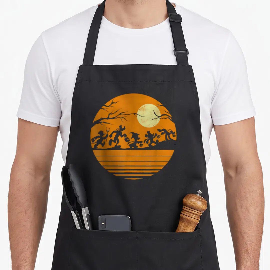 Friend halloween apron - Circular orange graphic with grey silhouettes, pale yellow moon, tree branches, and horizontal — shown on a black cotton canvas apron against a white background